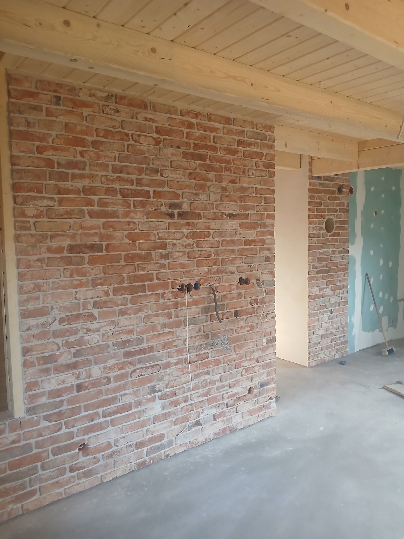 Ściana z czerwonej cegły dekoracyjnej w surowym wnętrzu z drewnianym stropem. Widoczne przewody elektryczne i fragment ściany z płyt kartonowo-gipsowych.