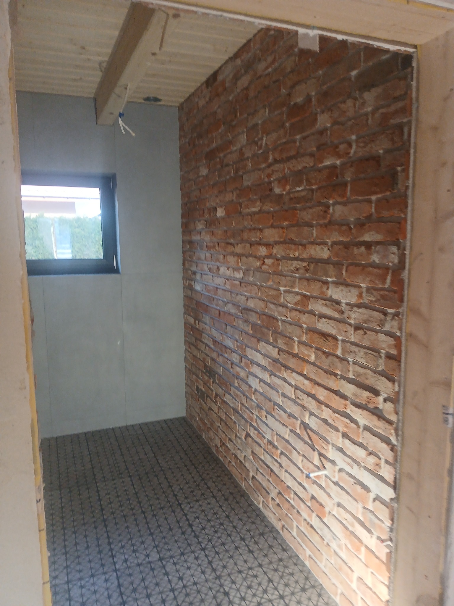 Wnętrze łazienki w trakcie wykończenia: ceglana ściana, szare płytki na ścianie z oknem, siatka na podłodze przygotowana pod wylewkę. Widoczne elementy drewniane konstrukcji.