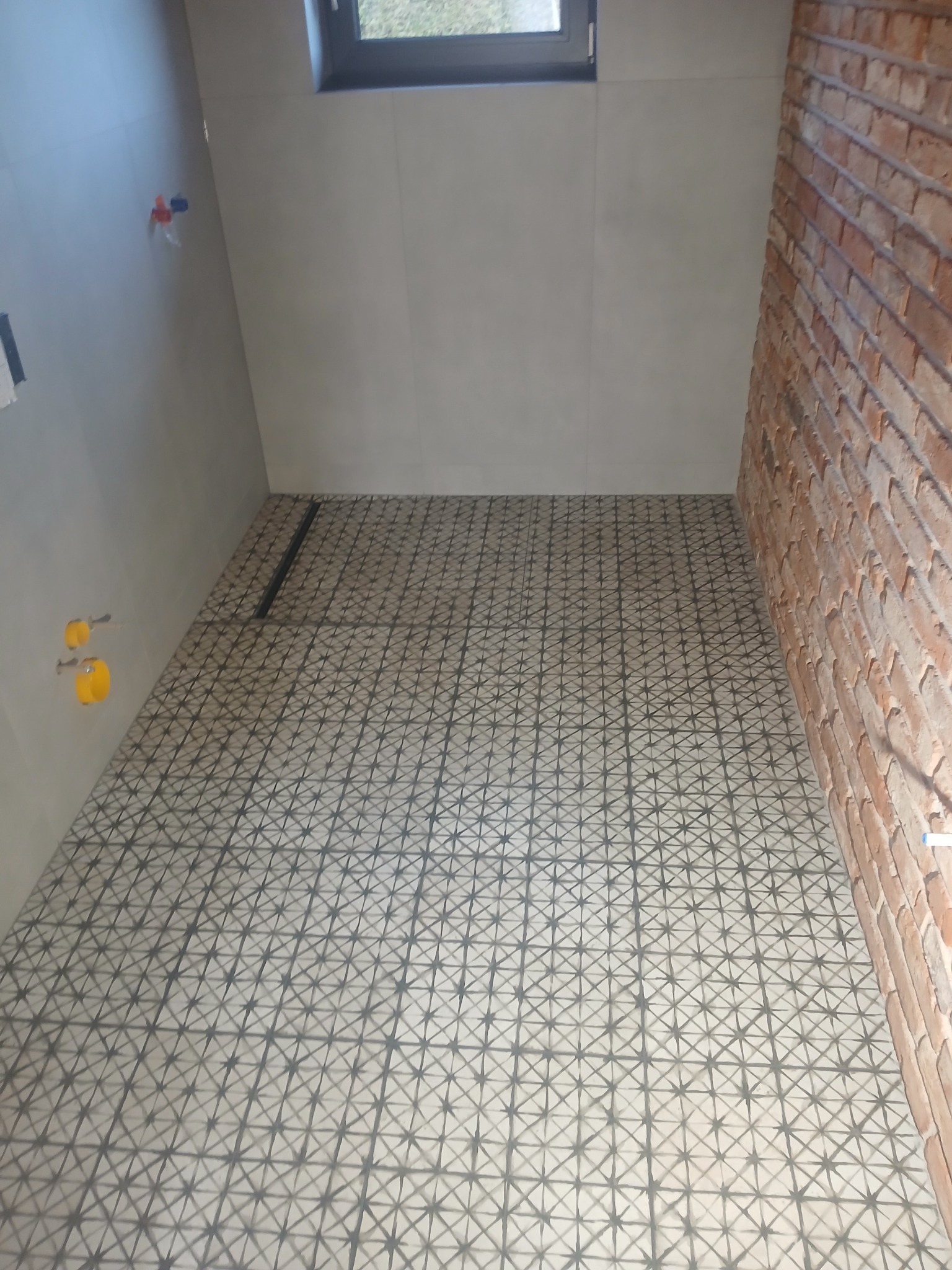 Wykończona łazienka z wzorzystą, szarą podłogą z odpływem liniowym, jasnymi płytkami ściennymi i ścianą z cegły. Widoczne elementy instalacji wodnej.