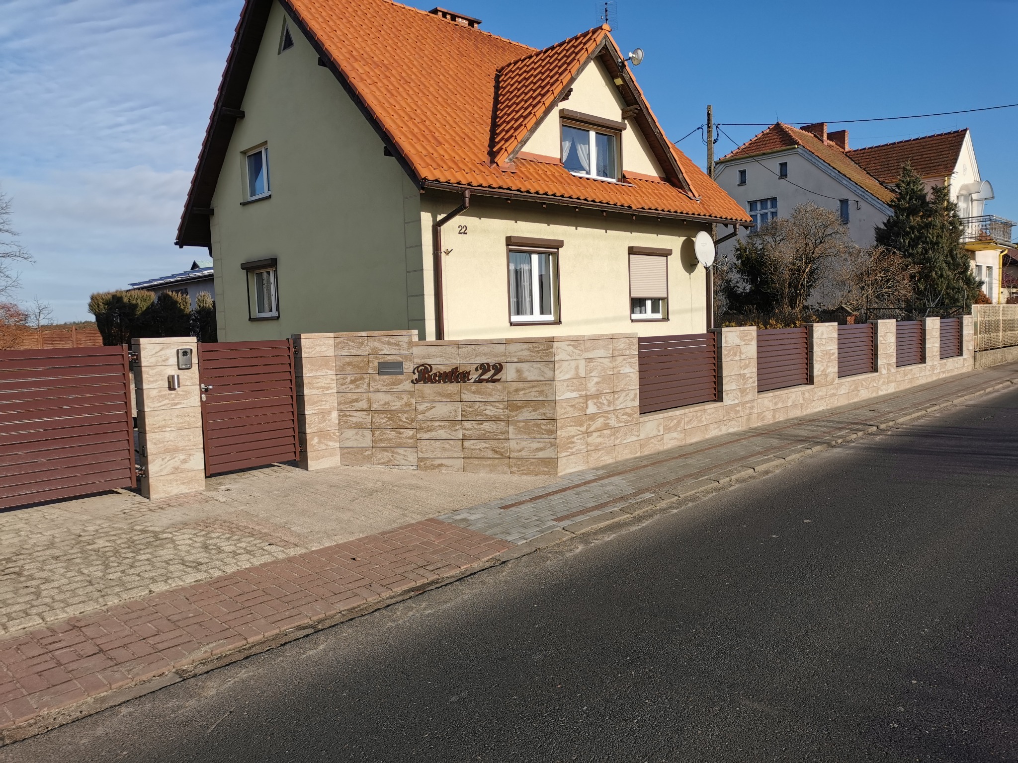 Nowoczesne ogrodzenie panelowe z brązowych, poziomych desek i beżowych, kwadratowych bloczków otaczające dom z numerem 22, widok z ulicy w słoneczny dzień.