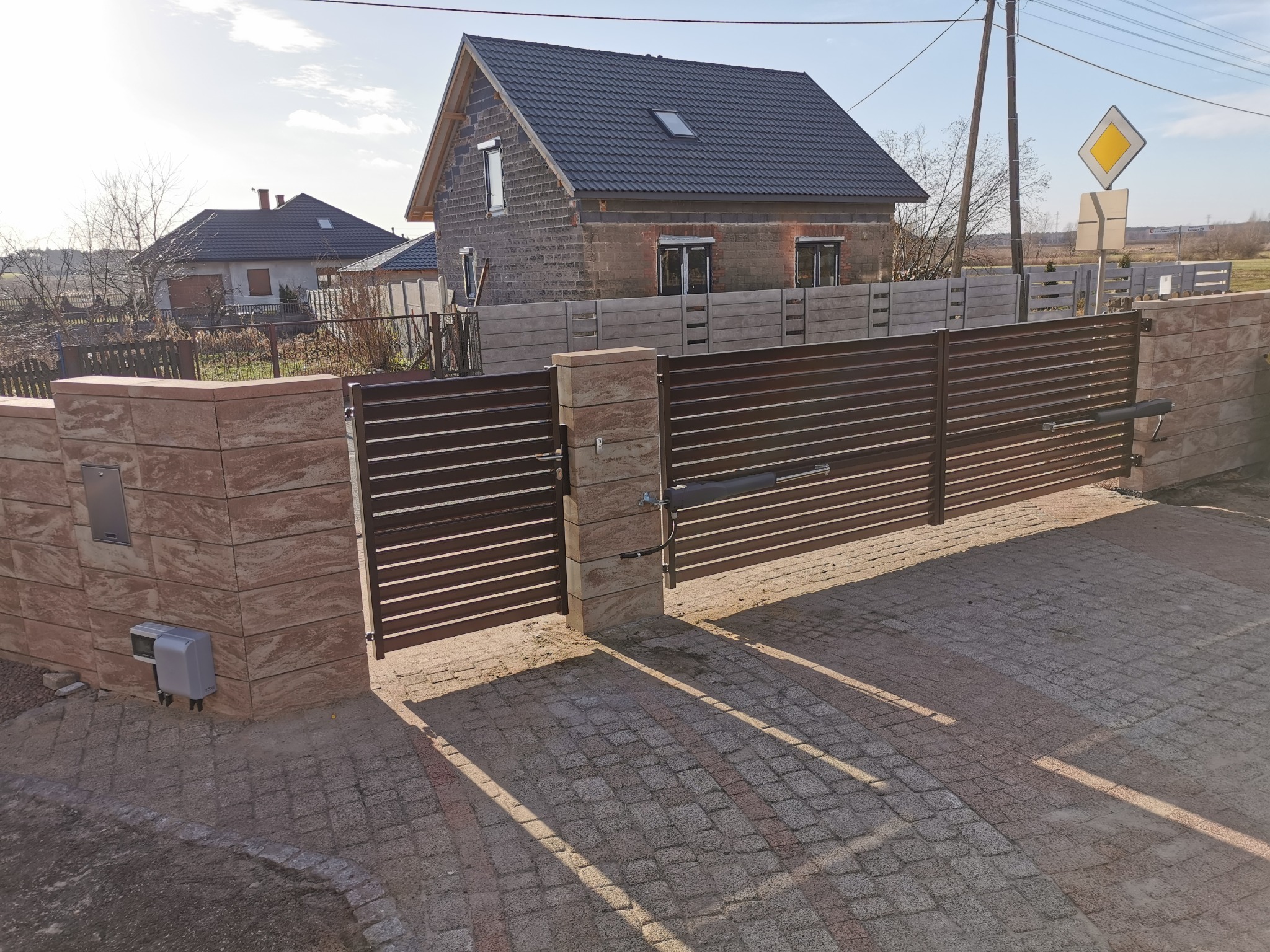 Automatyczna brama przesuwna z poziomymi panelami w kolorze ciemnego brązu, zamontowana między kamiennymi słupkami z jasnego piaskowca, na tle domu w trakcie budowy i kostki brukowej.