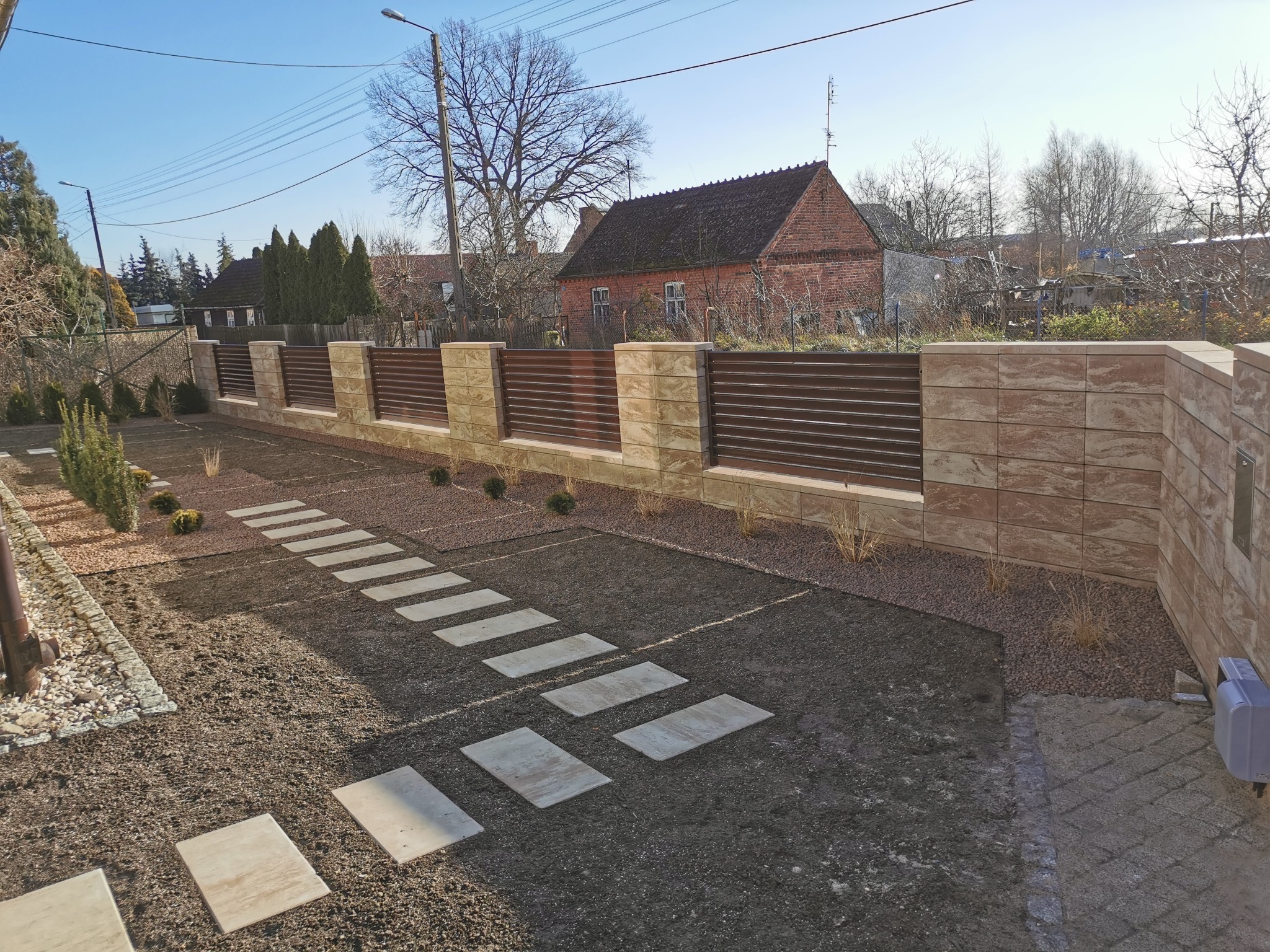 Nowoczesne ogrodzenie panelowe z poziomymi, brązowymi przęsłami i słupkami z beżowej, wzorzystej kostki, prowadzące do domu po ścieżce z płyt na świeżo usypanej ziemi.