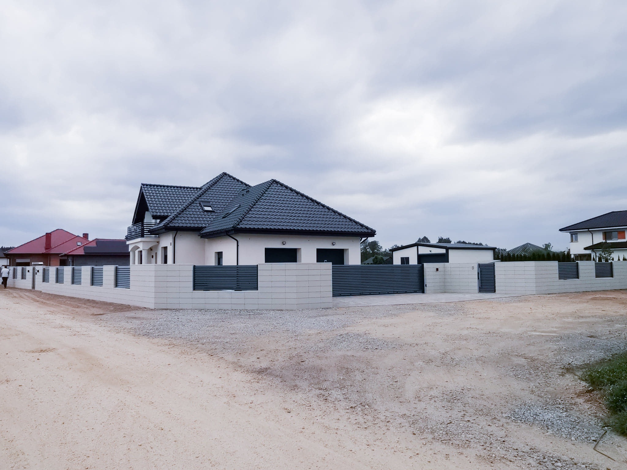 Nowoczesny dom jednorodzinny z szarą dachówką i jasną elewacją, otoczony ogrodzeniem z betonowych bloków i poziomych, grafitowych przęseł, widok z przodu na niezagospodarowaną działkę z piaskiem...