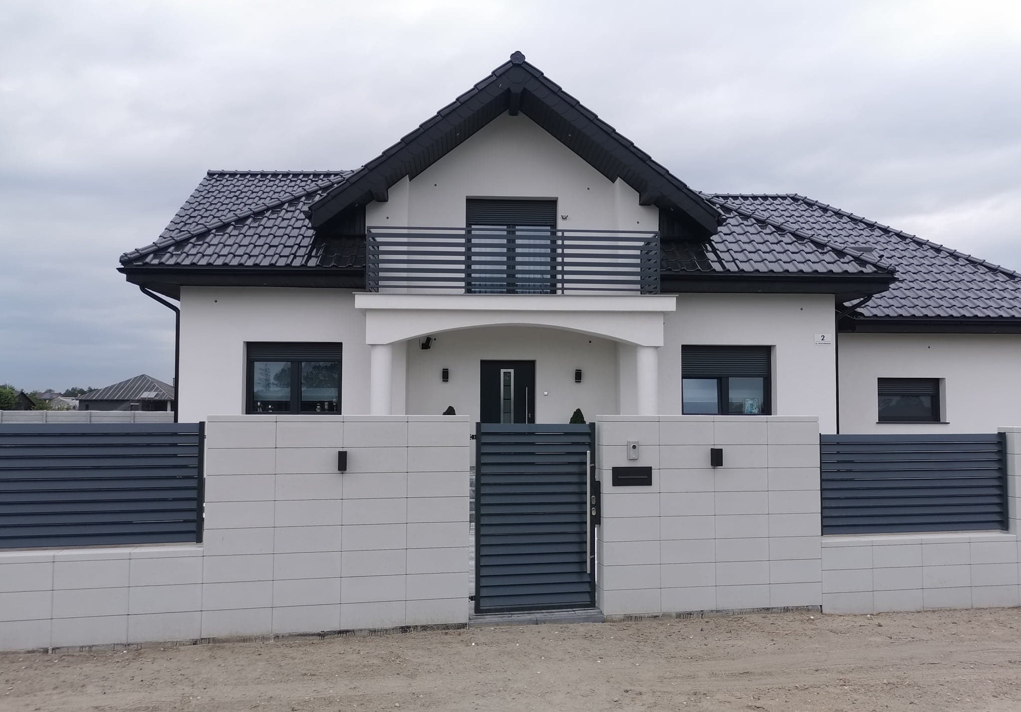 Nowoczesny dom jednorodzinny z białymi ścianami, ciemną dachówką i szarą, poziomą bramą wjazdową oraz ogrodzeniem z betonowych bloków, oświetlenie zewnętrzne.