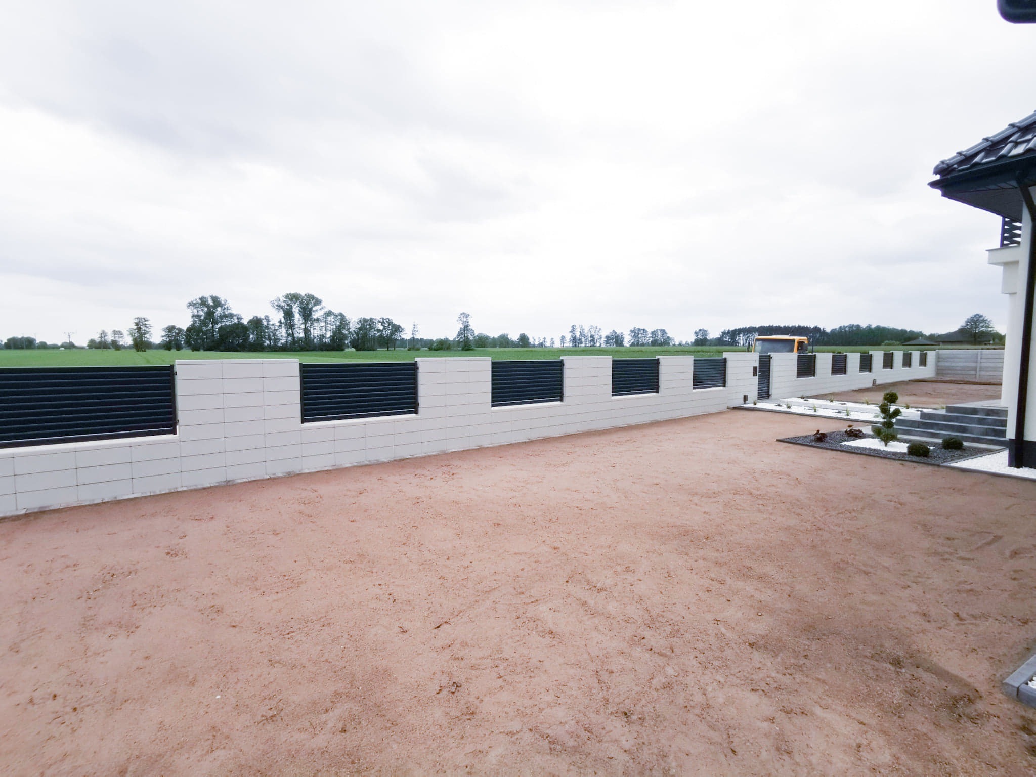 Nowoczesne ogrodzenie panelowe z elementami z jasnych płytek, widok na posesję z przygotowanym podłożem pod trawnik i fragment domu z czarnym dachem.