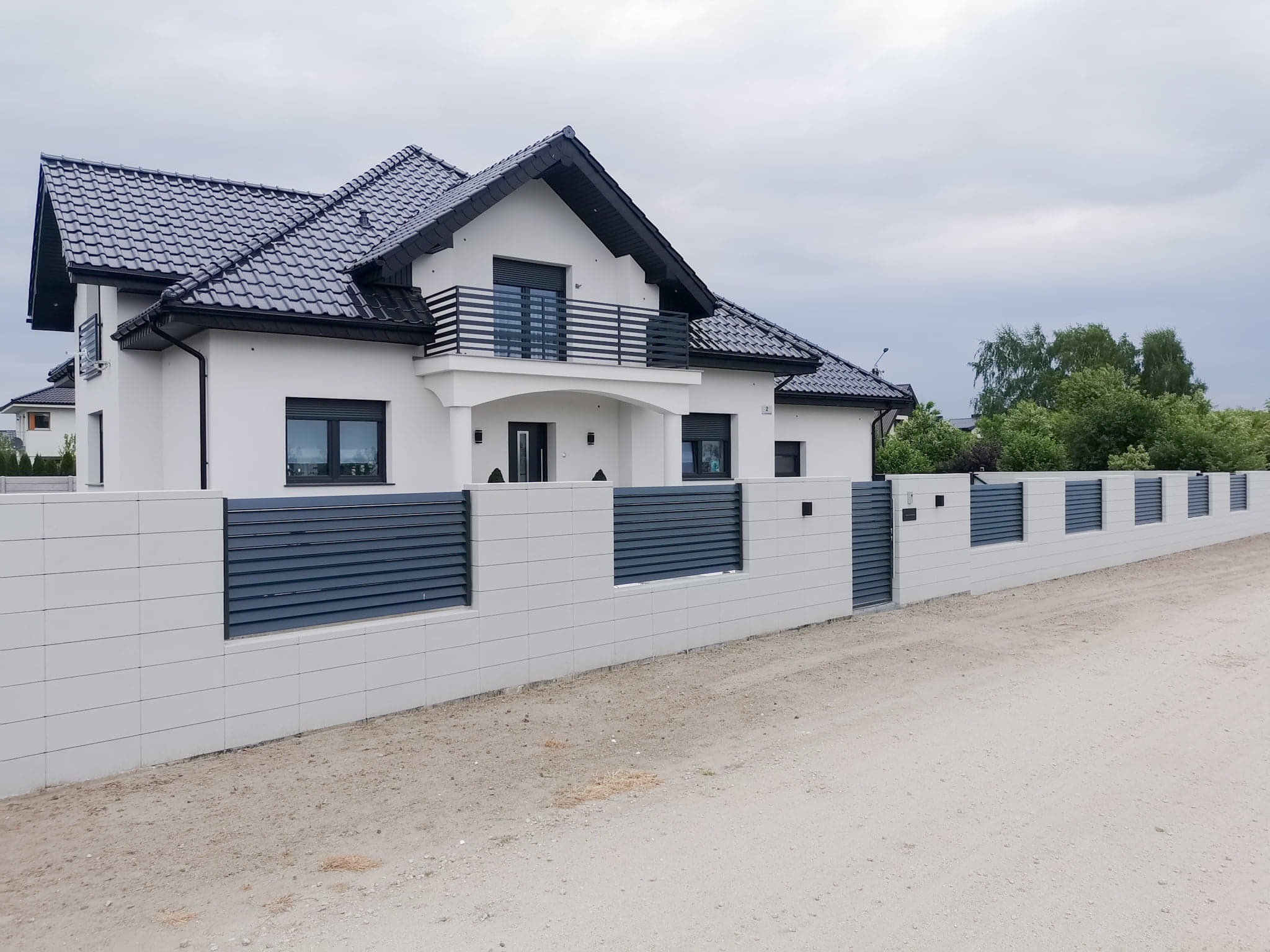 Nowoczesny, biały dom z ciemną dachówką i szarym, modułowym ogrodzeniem z poziomych paneli, widziany z poziomu gruntu.