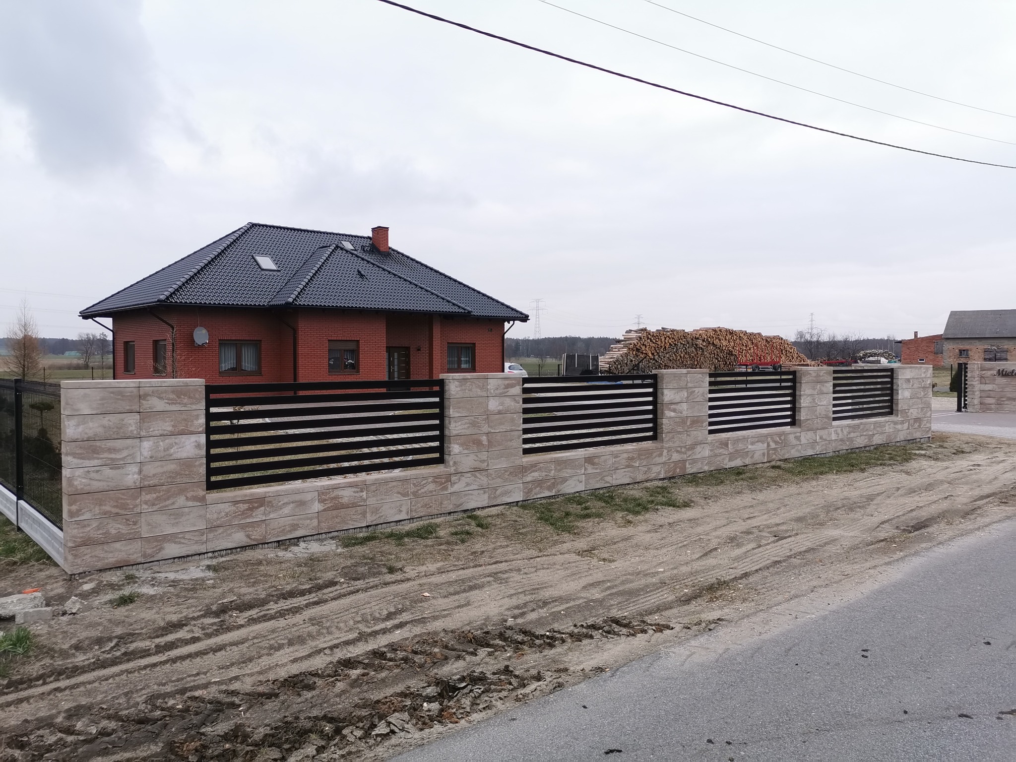 Nowoczesne ogrodzenie panelowe z elementami z kamienia dekoracyjnego, otaczające dom z czerwoną cegłą i ciemnym dachem, na tle pochmurnego nieba.