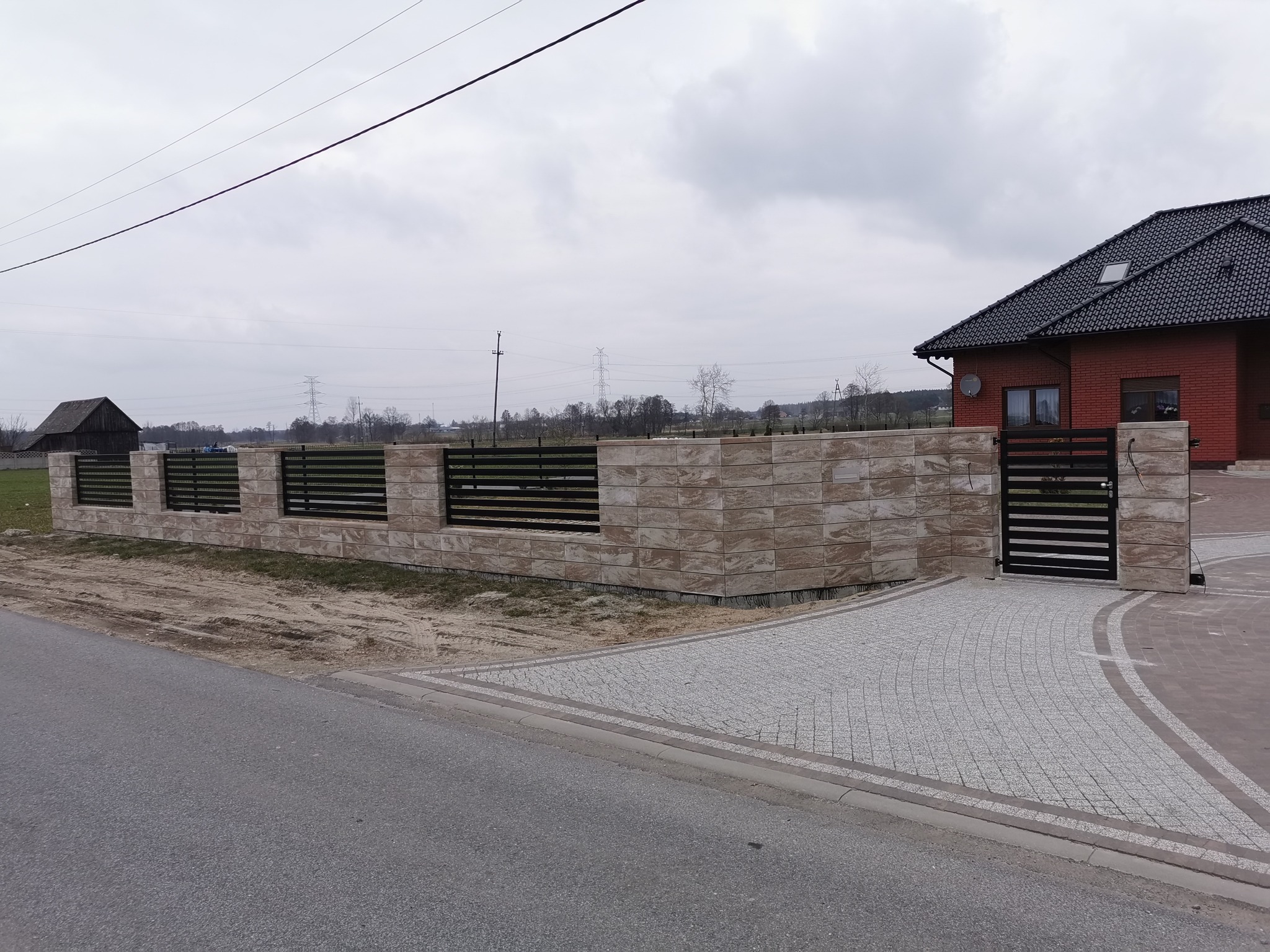 Nowoczesne ogrodzenie z elementami betonowymi i poziomymi przęsłami metalowymi, brama wjazdowa z tego samego materiału, posesja z kostką brukową, w tle dom z czerwonej cegły i szare niebo.