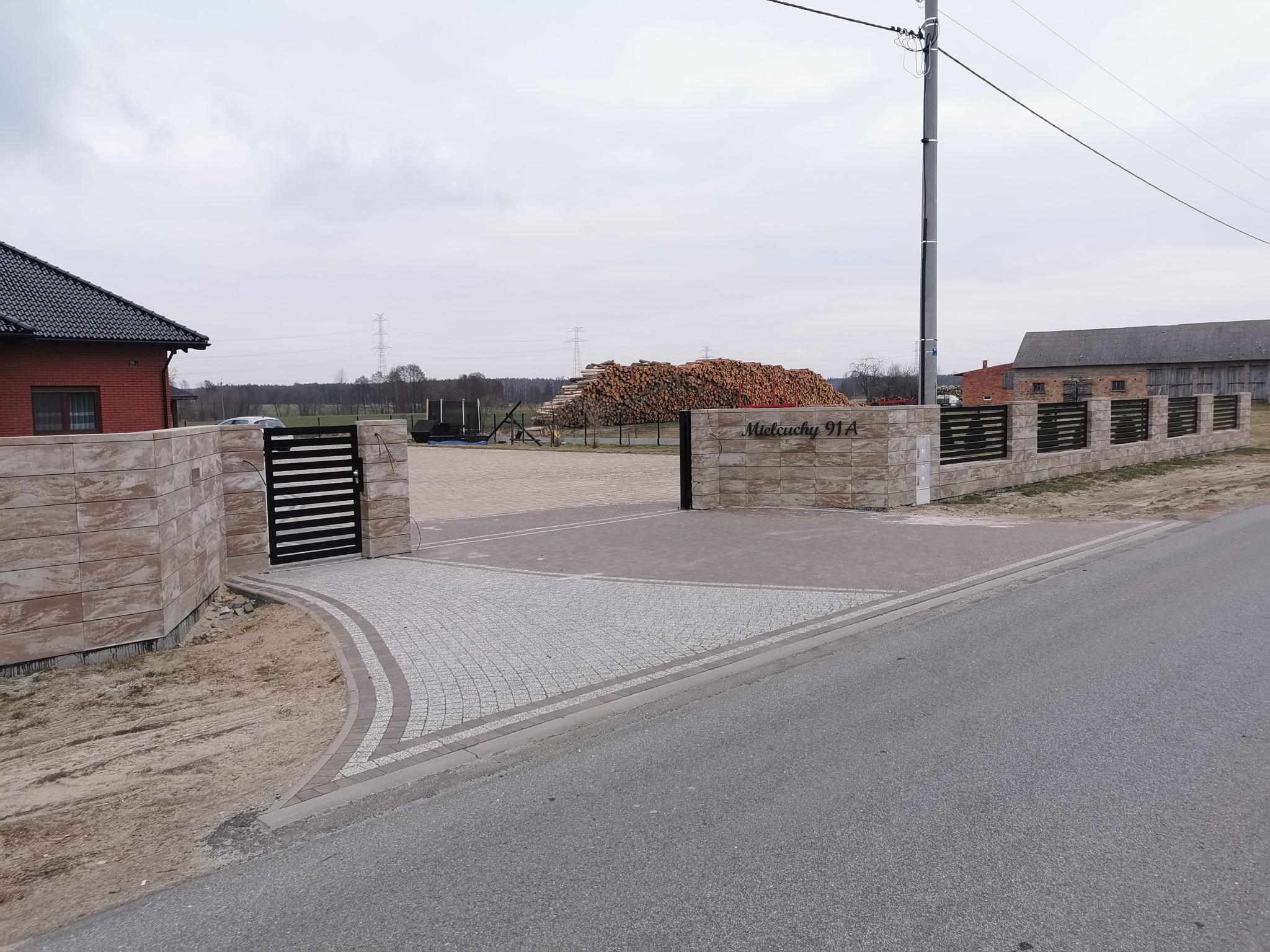 Nowoczesne ogrodzenie panelowe z bramą przesuwną i furtką, wykończone piaskowcem, z adresem 'Mielcuchy 91A', na tle sterty drewna i pochmurnego nieba.