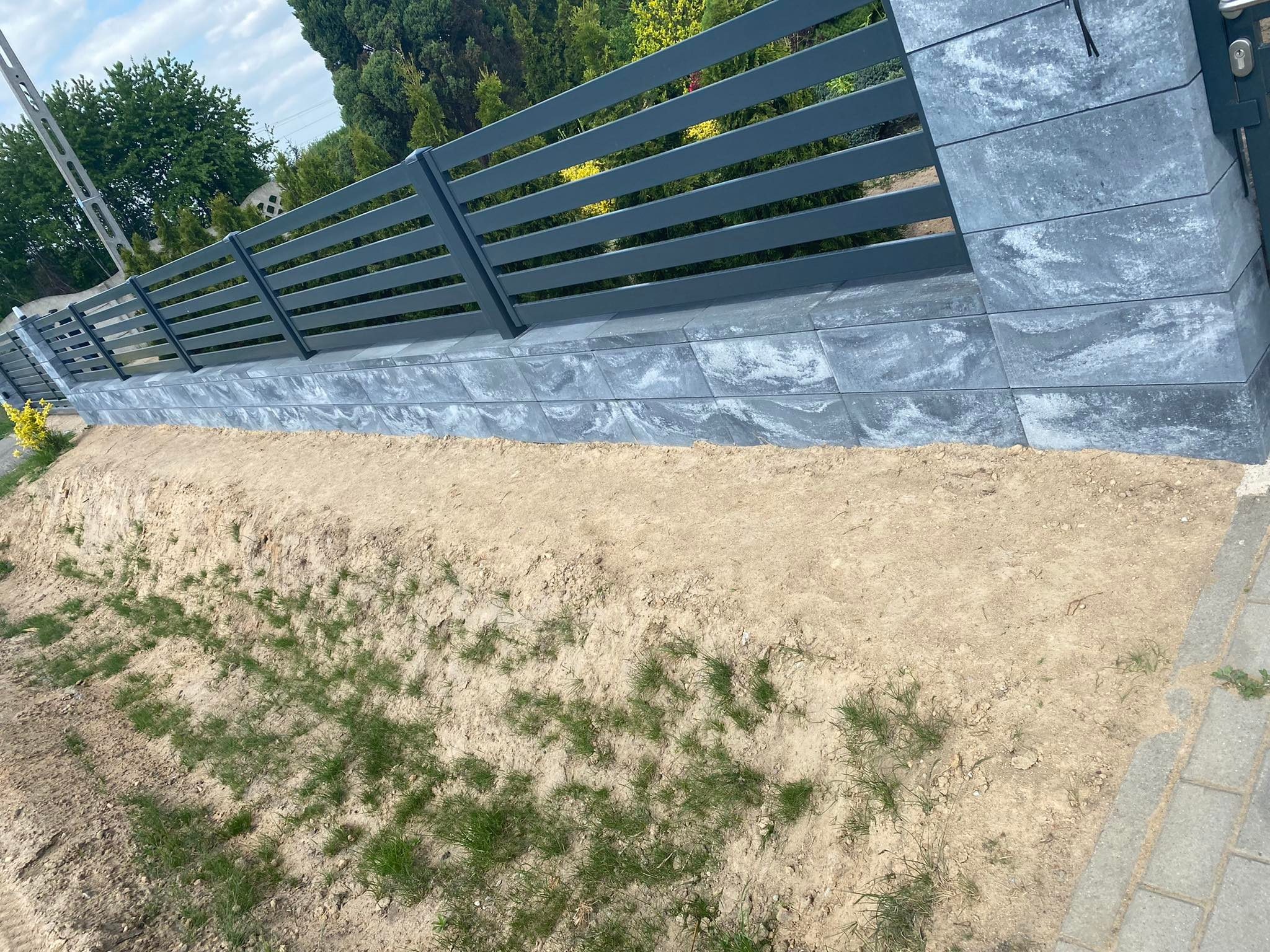 Nowoczesne ogrodzenie panelowe w kolorze grafitowym, z podmurówką z szarej kostki imitującej kamień, na tle zieleni drzew i krzewów. Widoczny fragment chodnika z kostki brukowej.
