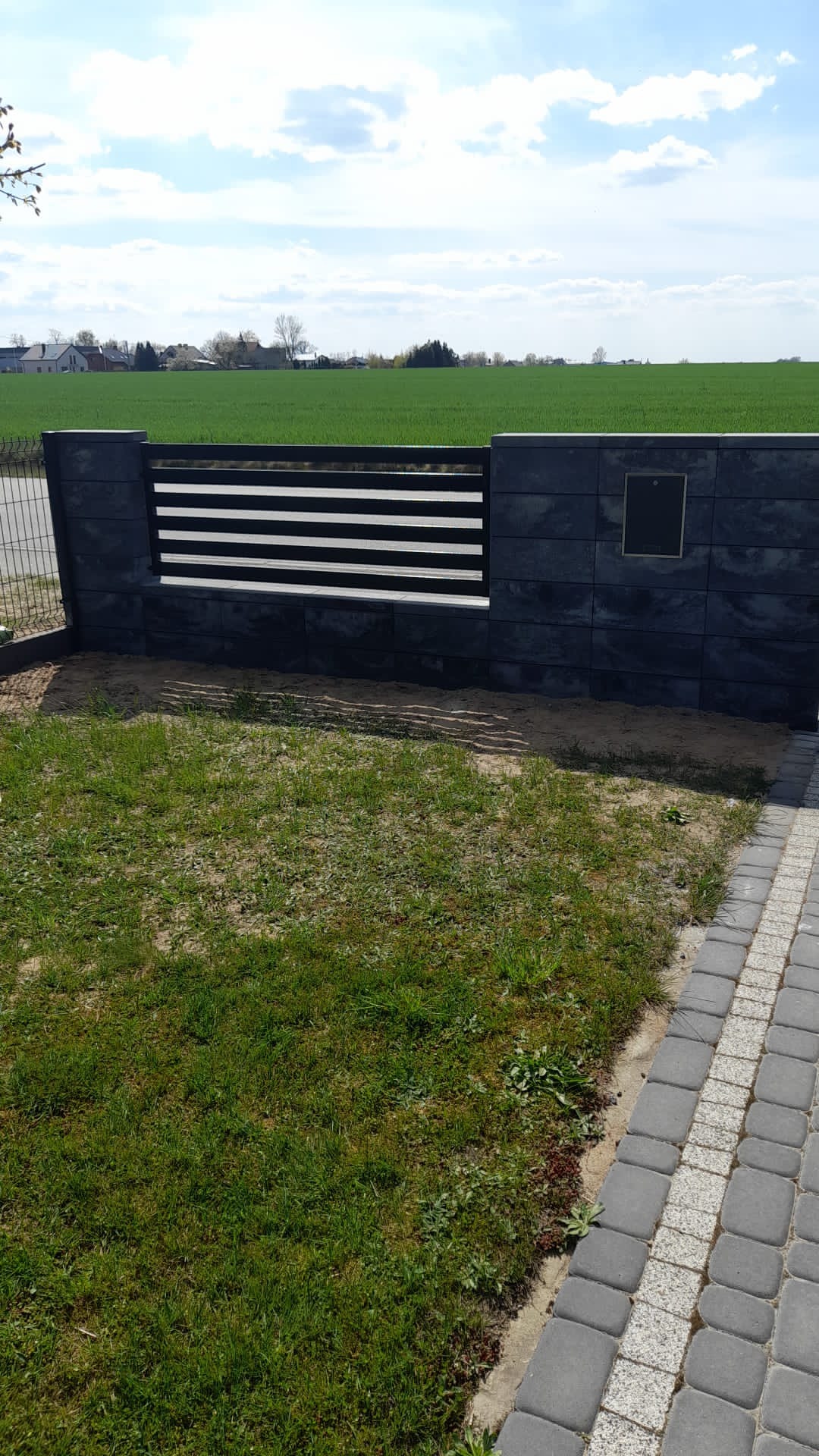 Nowoczesne ogrodzenie panelowe z poziomymi, czarnymi przęsłami, osadzone na słupkach z szarego betonu łupanego, z widoczną tabliczką adresową, na tle zielonego pola i błękitnego nieba.