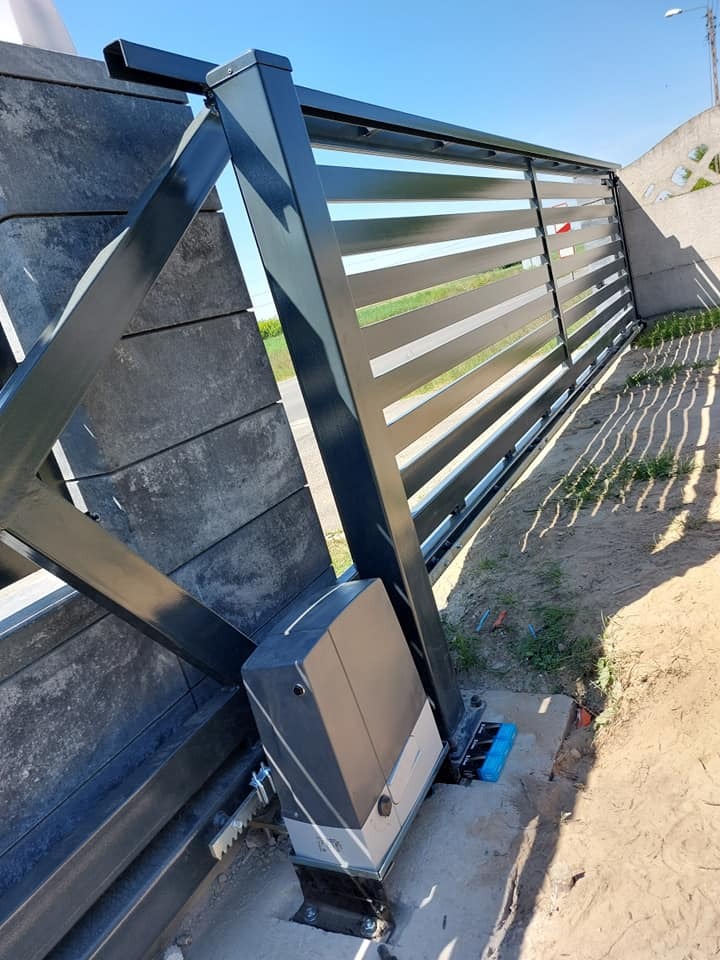 Szara, przesuwna brama z poziomymi panelami i automatycznym napędem zamontowanym na betonowej podstawie, widok z boku na tle niebieskiego nieba.