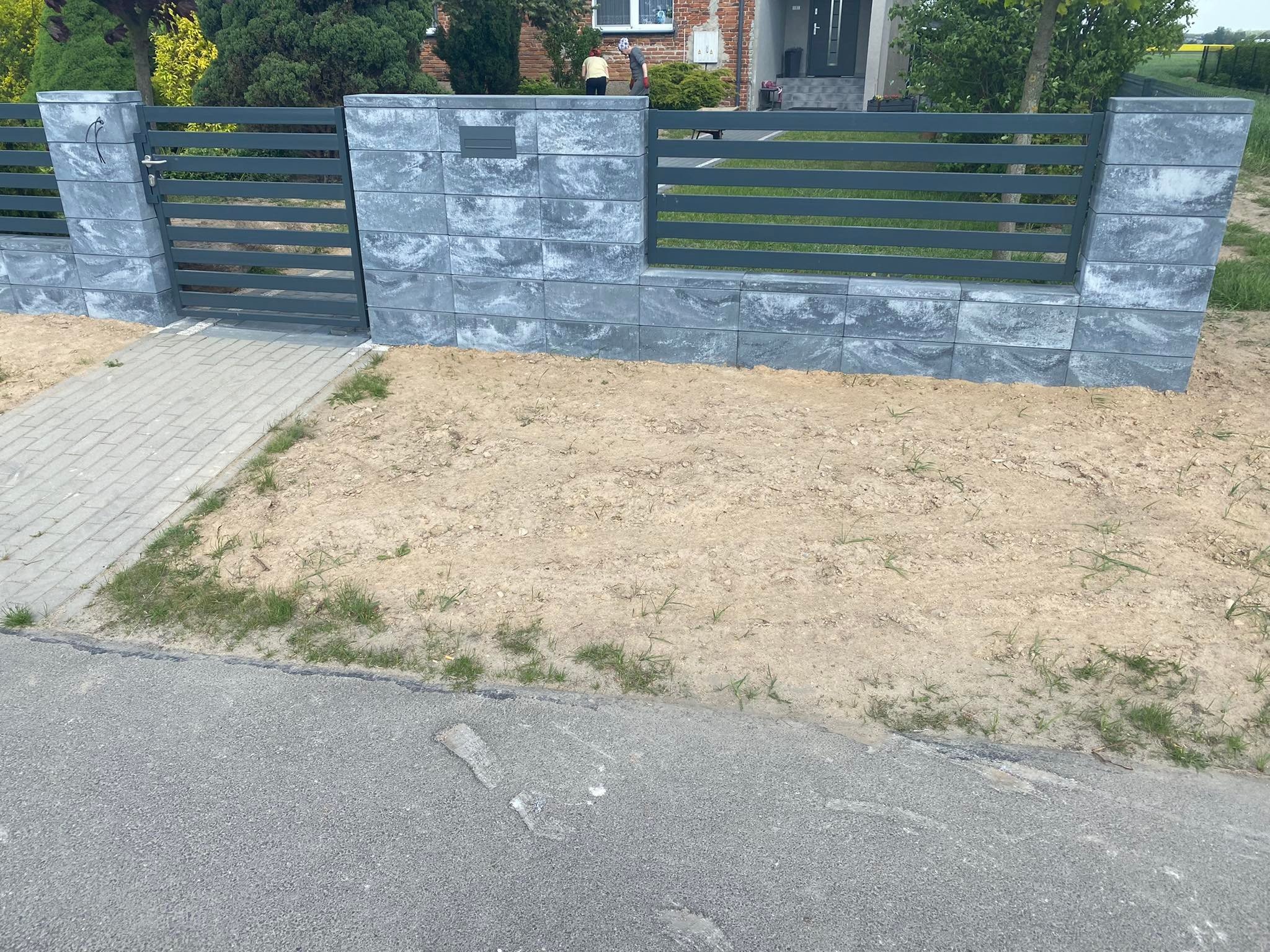 Nowoczesne ogrodzenie panelowe z szarej kostki betonowej i poziomych metalowych przęseł, furtka z klamką, widok na dom z cegły w tle, teren wokół ogrodzenia w trakcie prac ziemnych.