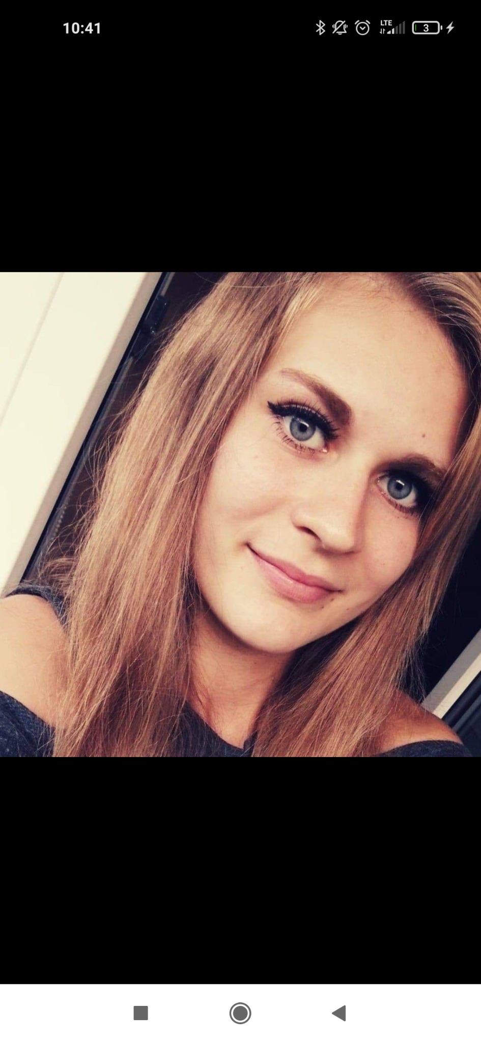 Portret młodej kobiety z blond włosami i niebieskimi oczami, skierowanej w stronę obiektywu, z delikatnym uśmiechem.