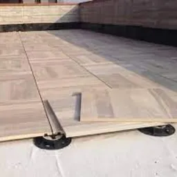 Montaż desek tarasowych na regulowanych wspornikach na białej membranie izolacyjnej, widoczne szczeliny dylatacyjne i fragment ściany w tle.