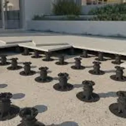 Montaż tarasu na regulowanych wspornikach, widoczne płyty tarasowe częściowo ułożone na wspornikach, piasek pod płytami, budynek w tle.