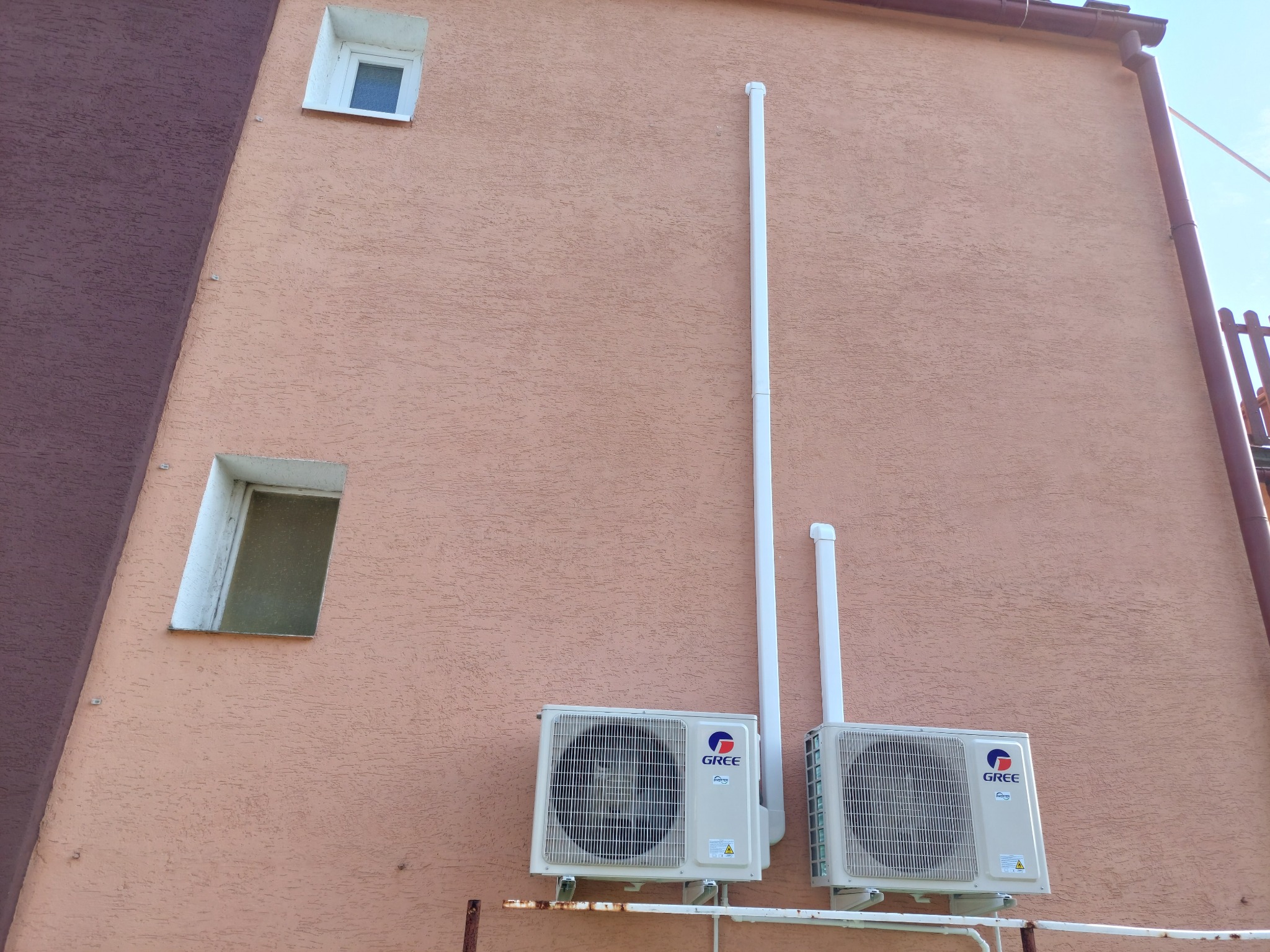 Dwa zewnętrzne agregaty klimatyzacji marki Gree zamontowane na ścianie budynku z oknami, widoczne białe rury odprowadzające skropliny.