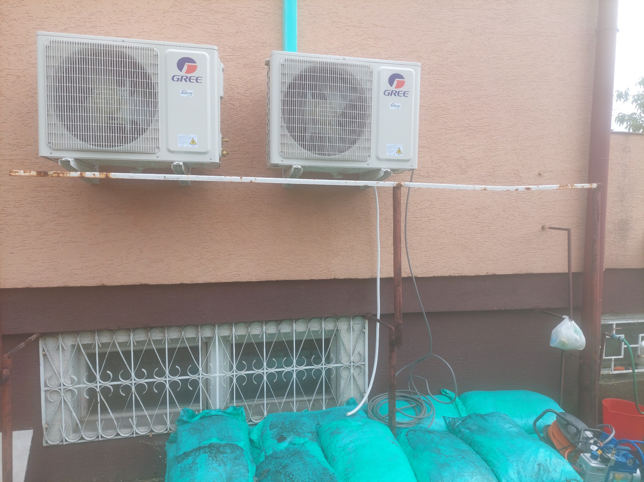 Dwa klimatyzatory marki Gree zamontowane na ścianie budynku, z widocznymi przewodami i workami z materiałem izolacyjnym u dołu.