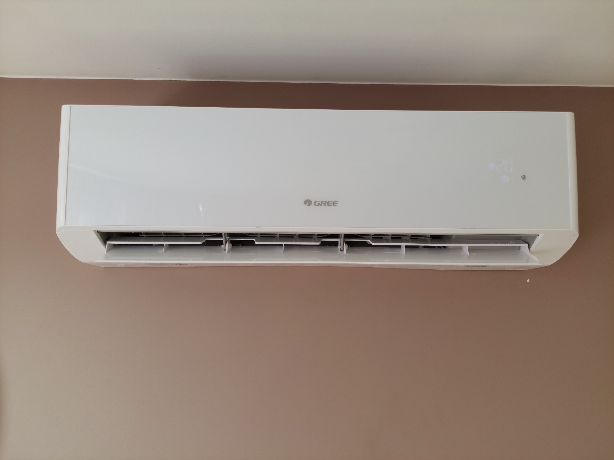 Biała jednostka klimatyzacyjna marki Gree zamontowana na ścianie o kolorze kawy z mlekiem, wyświetlająca temperaturę 20 stopni Celsjusza.
