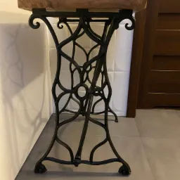 Stolik z blatem z naturalnego drewna na ozdobnej, metalowej podstawie w stylu retro, na tle ściany z białych płytek i brązowych drzwi.