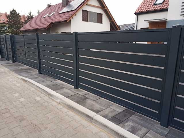 Nowoczesne, poziome, grafitowe przęsła ogrodzeniowe z metalu, widoczne od strony chodnika z kostki brukowej, z domami w tle.