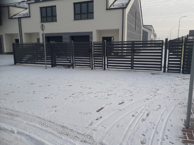 Nowoczesna, pozioma, metalowa brama wjazdowa i przęsła ogrodzenia w kolorze antracytowym, przed nowym budynkiem mieszkalnym, pokryte śniegiem, widoczne ślady opon i stóp na śniegu.
