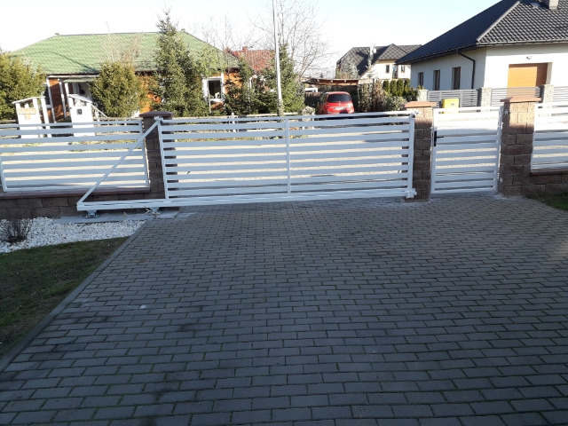 Automatyczna brama przesuwna, biała, z poziomymi panelami, zamontowana na murku z cegły klinkierowej, widok na posesję z kostki brukowej i domy w tle.