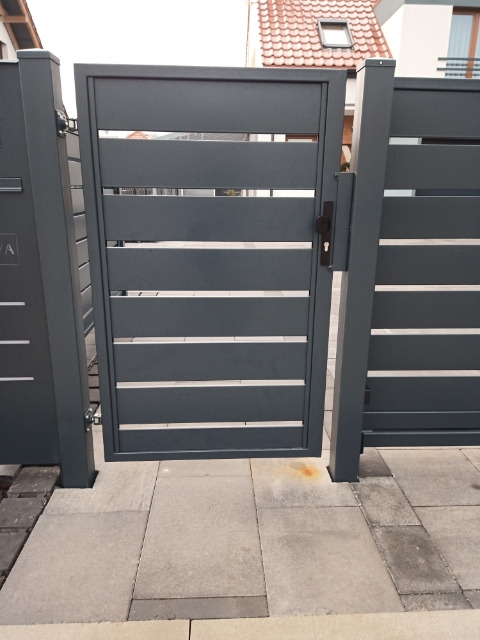 Nowoczesna, grafitowa furtka z poziomymi panelami, z czarną klamką i zamkiem, osadzona w słupkach na jasnoszarej kostce brukowej, z widocznym fragmentem domu w tle.
