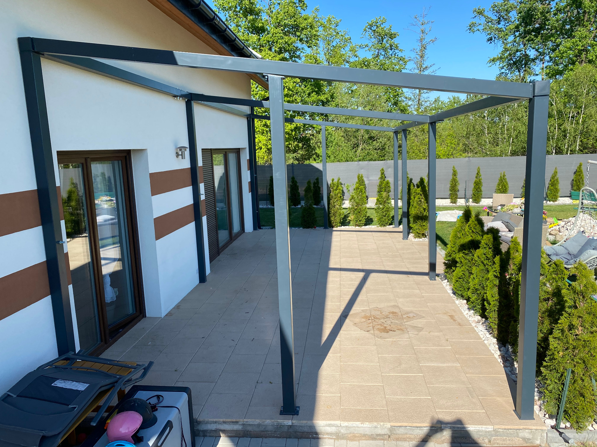 Nowoczesna, szara pergola metalowa na tarasie z jasnej kostki brukowej, widok na ogród z tujami i szarym ogrodzeniem panelowym w tle.