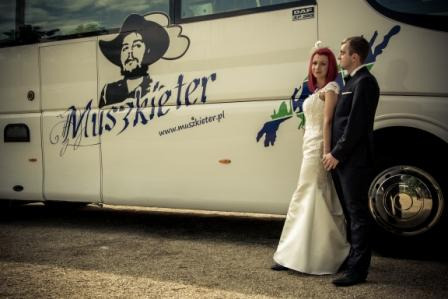 Para młoda w strojach ślubnych pozuje przed autobusem z logo firmy przewozowej 'Muszkieter' z grafiką muszkietera w kapeluszu.
