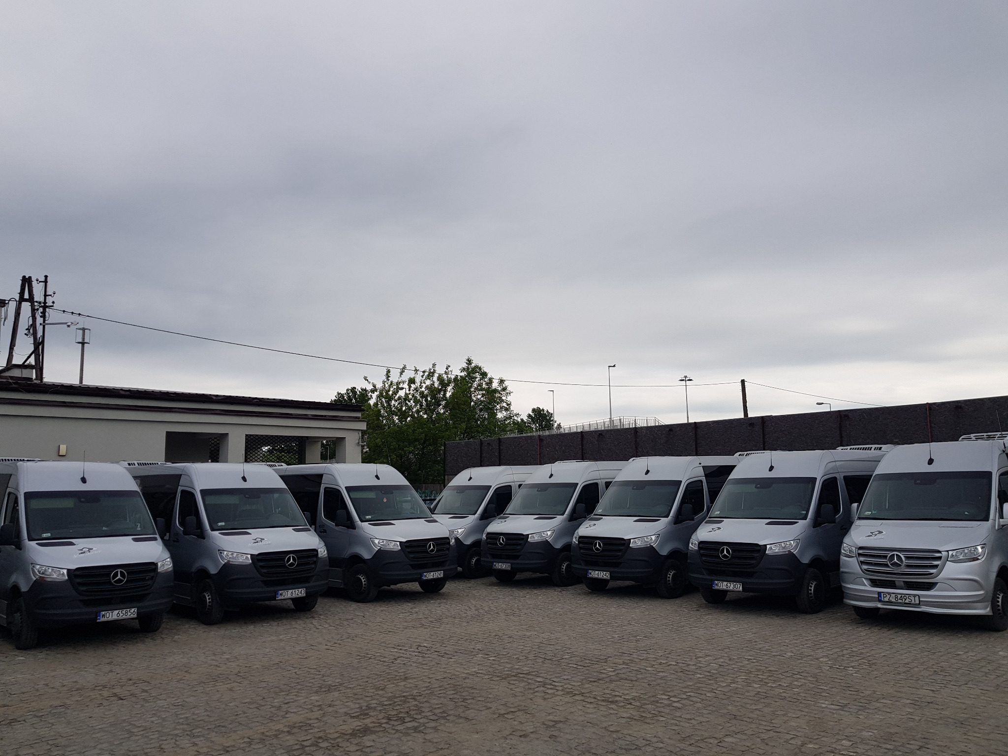 Rząd srebrnych busów Mercedes-Benz Sprinter zaparkowanych na brukowanym placu, widok z przodu, pochmurne niebo w tle.