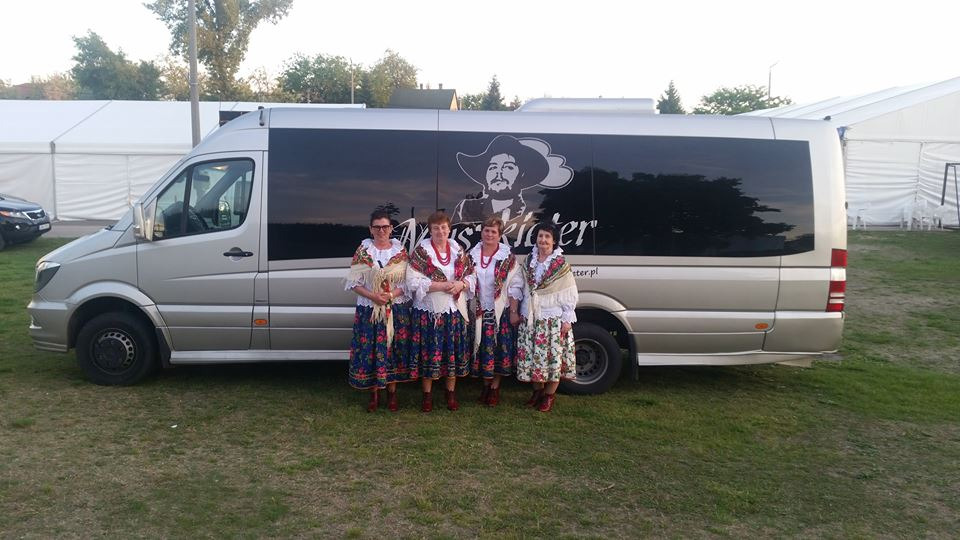 Cztery kobiety w strojach ludowych stoją przed srebrnym busem z grafiką mężczyzny w kapeluszu i nazwą firmy 'Musketeer' na boku, na tle białego namiotu.