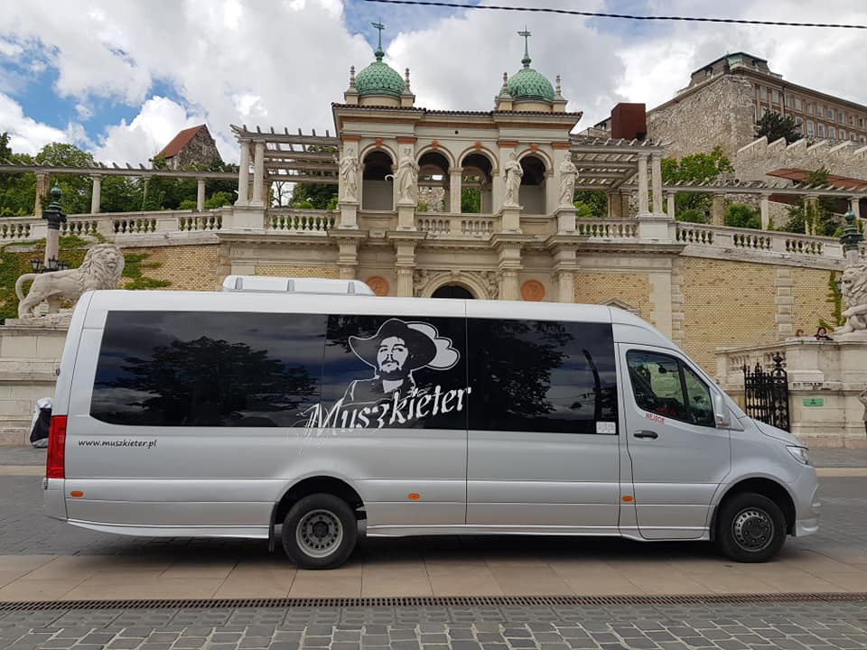 Srebrny bus z logo 'Muszkieter' na tle zabytkowej architektury z rzeźbami lwów w Budapeszcie.