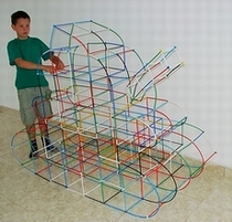 Chłopiec w szarej koszulce opiera ręce na dużej, przestrzennej konstrukcji z kolorowych rurek, przypominającej abstrakcyjny pojazd lub budowlę. Konstrukcja stoi na podłodze z płytek w jasnym...