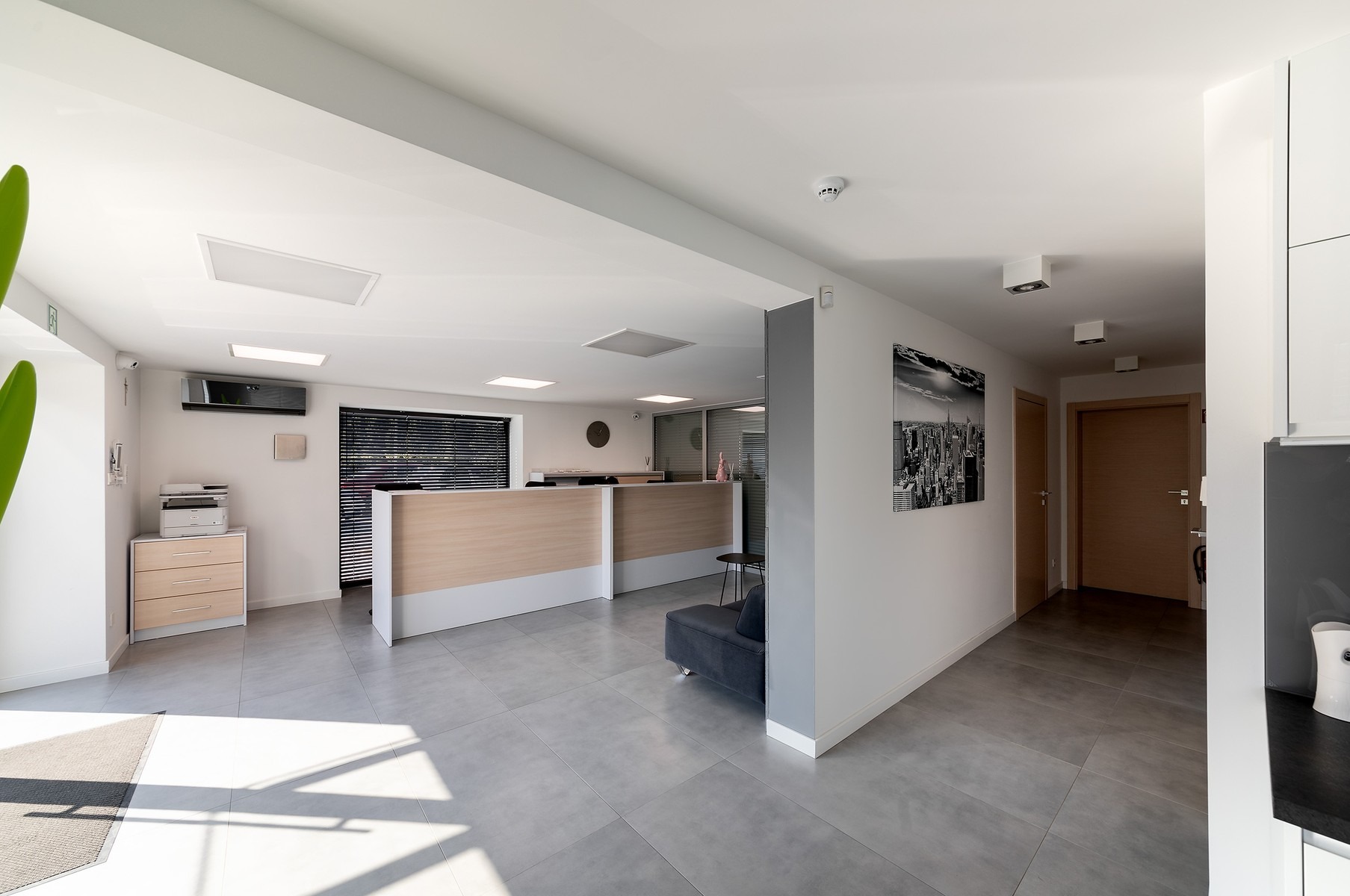 Jasne, przestronne biuro z recepcją o drewnianym wykończeniu, szarą podłogą i minimalistycznym wystrojem, widok na korytarz z czarno-białym zdjęciem miasta na ścianie.