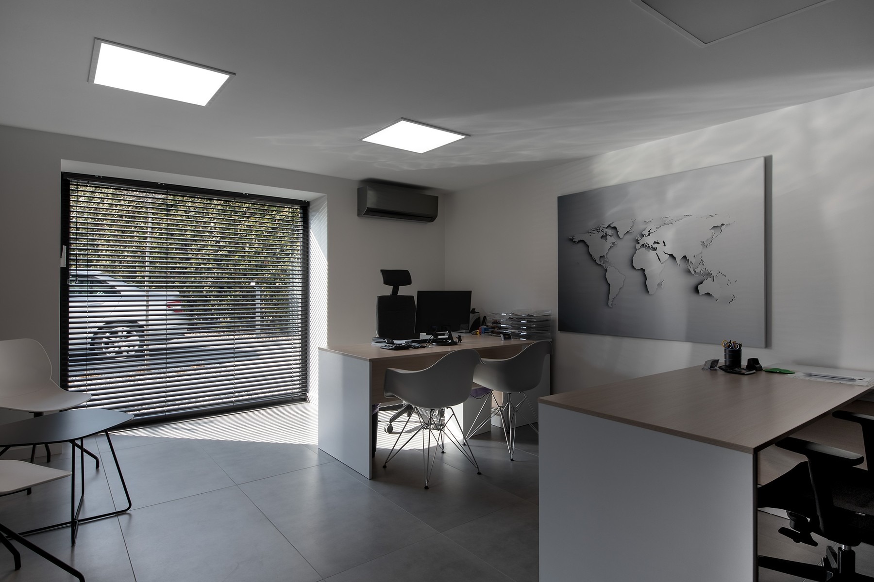 Jasne, minimalistyczne biuro w Pcimiu z dwoma biurkami, krzesłami i dużym oknem z żaluzjami. Na ścianie mapa świata, a na biurkach akcesoria biurowe i dokumenty.