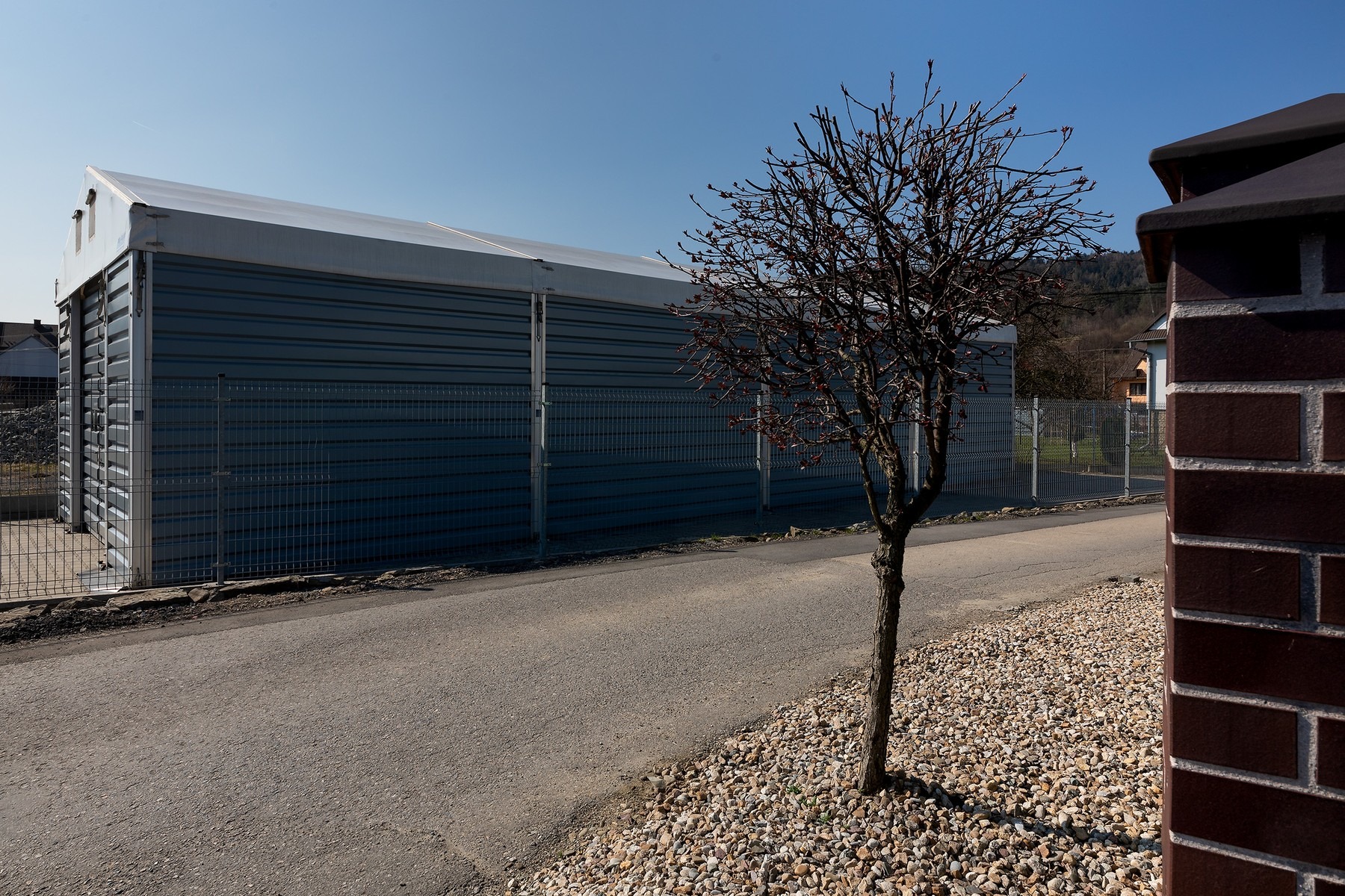 Szara hala magazynowa z metalowymi panelami i dachem z plandeki, ogrodzona siatką, widziana z poziomu ulicy z młodym drzewem z przodu i fragmentem ceglanego muru po prawej stronie.
