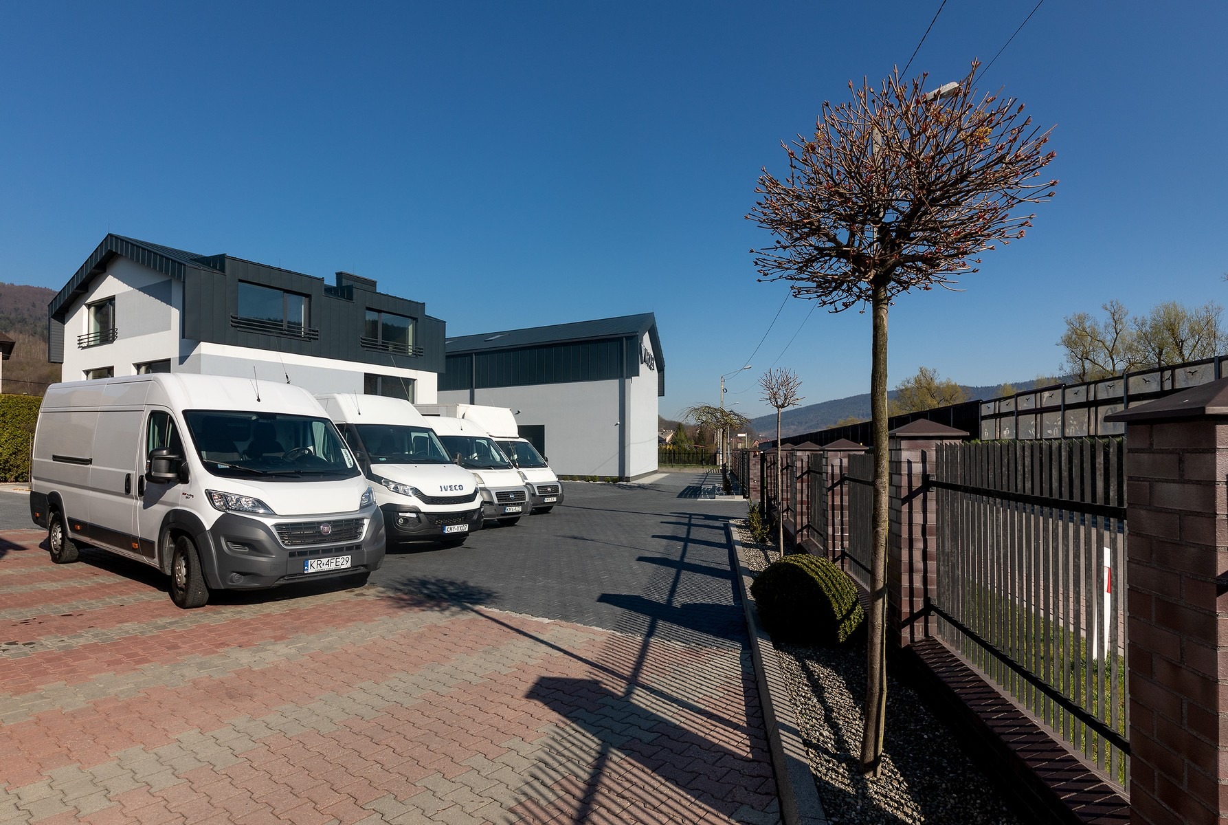 Flota białych samochodów dostawczych Iveco i Fiat na parkingu przed nowoczesnym budynkiem firmy, w tle błękitne niebo i drzewo bez liści.