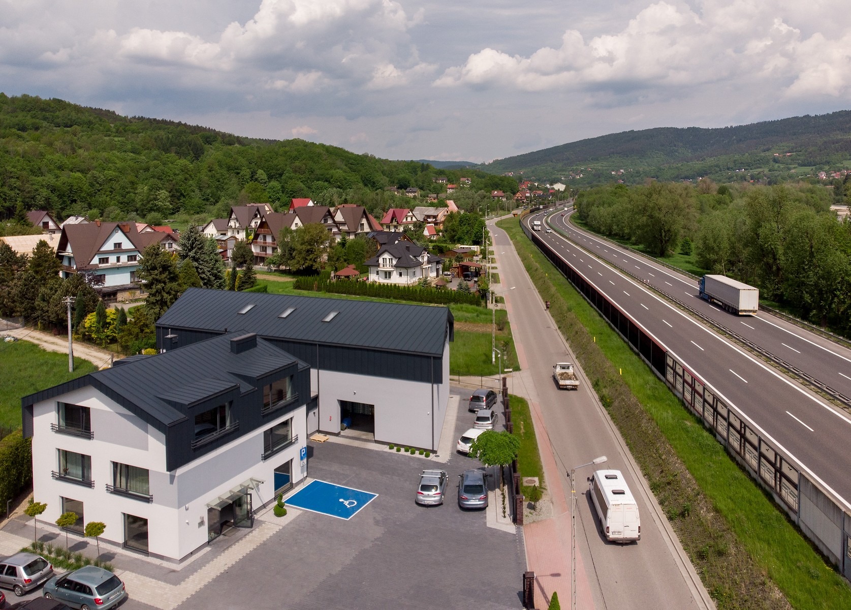Widok z góry na nowoczesny budynek biurowy z szarym dachem, zaparkowanymi samochodami i niebieskim znakiem parkingu dla niepełnosprawnych, w tle krajobraz z autostradą i zabudowaniami mieszkalnymi.