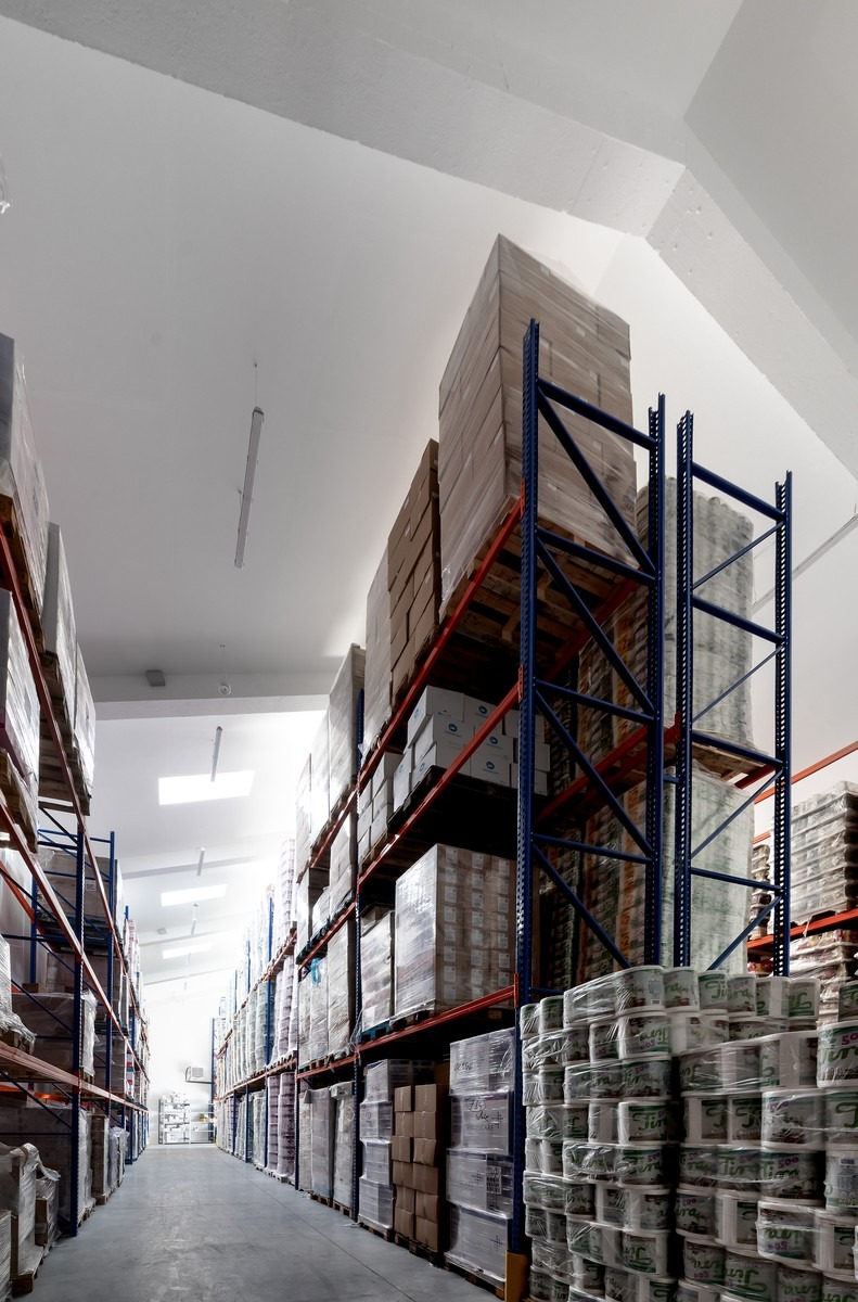 Wnętrze magazynu z wysokimi regałami wypełnionymi kartonami, pojemnikami i opakowaniami, oświetlonymi świetlikami w suficie.