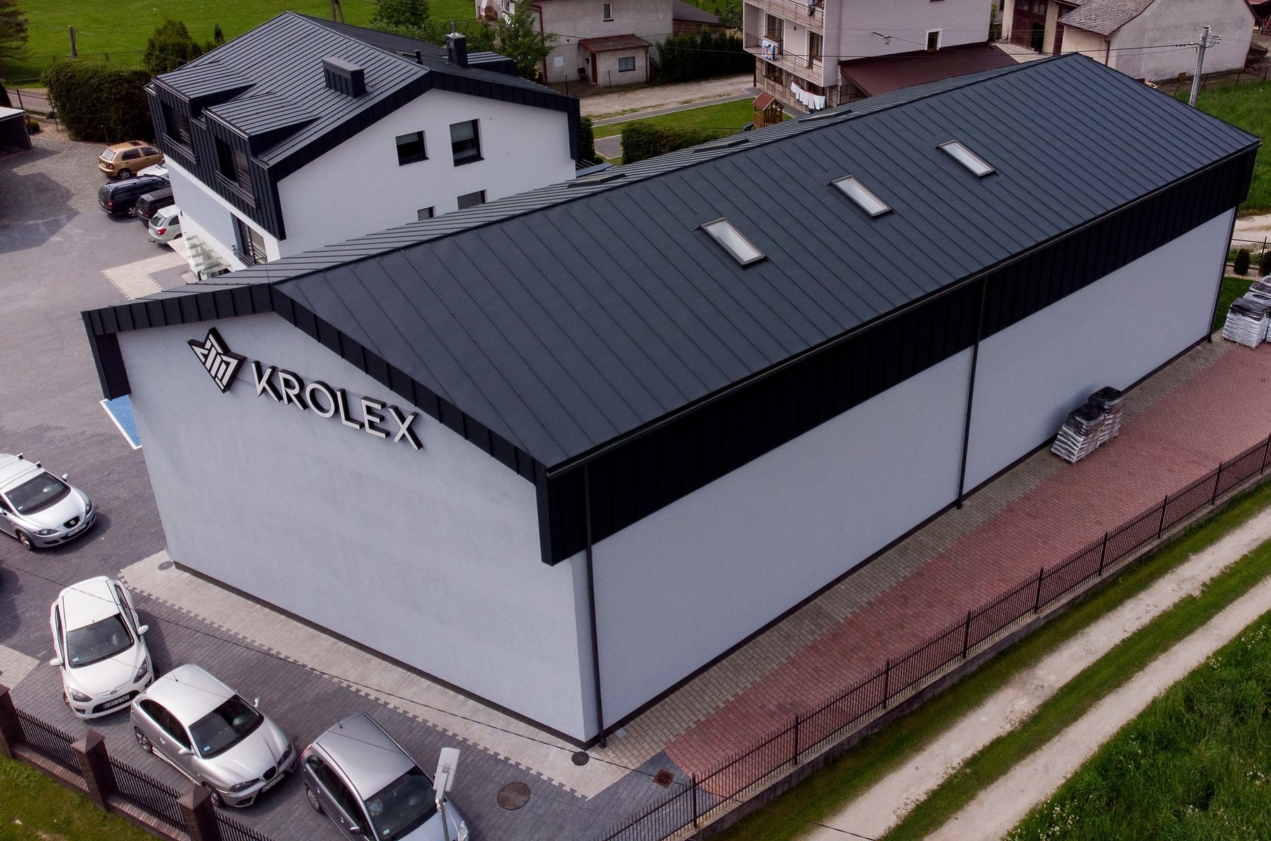 Budynek firmy Krolex z ciemnoszarą, metalową blachodachówką z trzema oknami dachowymi, widok z góry, na parkingu zaparkowane samochody, w tle zabudowa jednorodzinna.