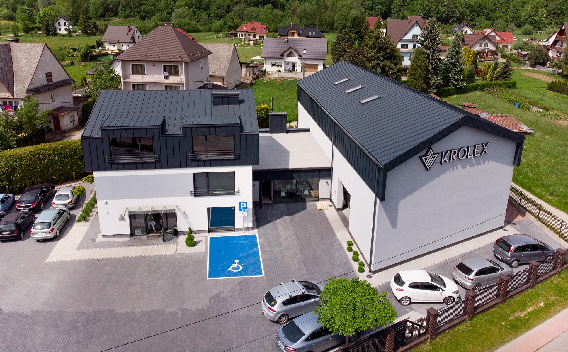 Widok z góry na nowoczesny budynek biurowy firmy Krolex z parkingiem, otoczony zielenią i zabudową jednorodzinną.