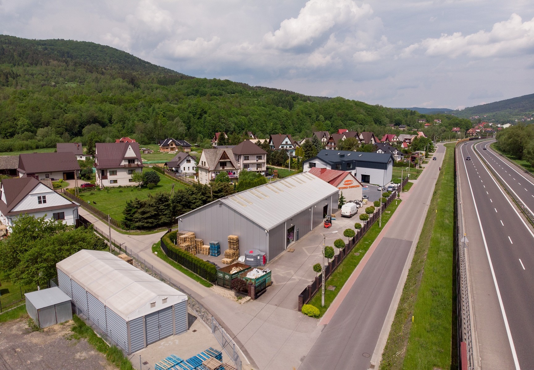 Widok z góry na magazyn z paletami, kontenerem i wózkiem widłowym na tle górskiego krajobrazu i autostrady.