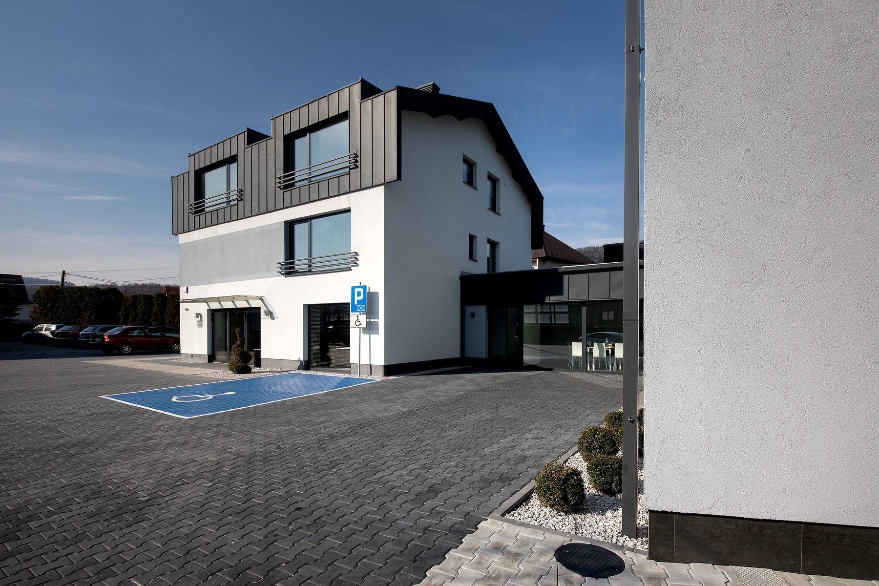 Nowoczesny budynek biurowy z parkingiem wyłożonym kostką brukową, oznaczonym miejscem dla niepełnosprawnych i schludną zielenią.