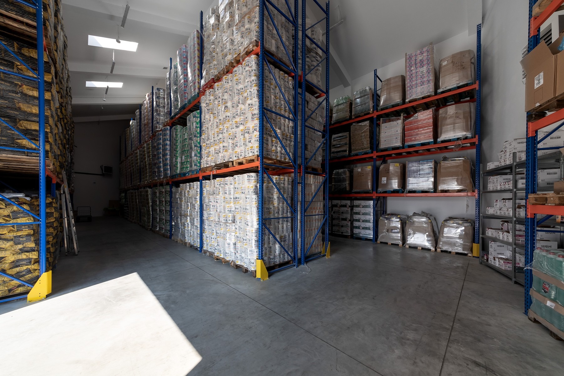 Magazyn z wysokimi regałami wypełnionymi zapasami, w tym paletami z opakowaniami papieru toaletowego i workami z proszkiem do prania, oświetlony światłem dziennym wpadającym przez okna dachowe.