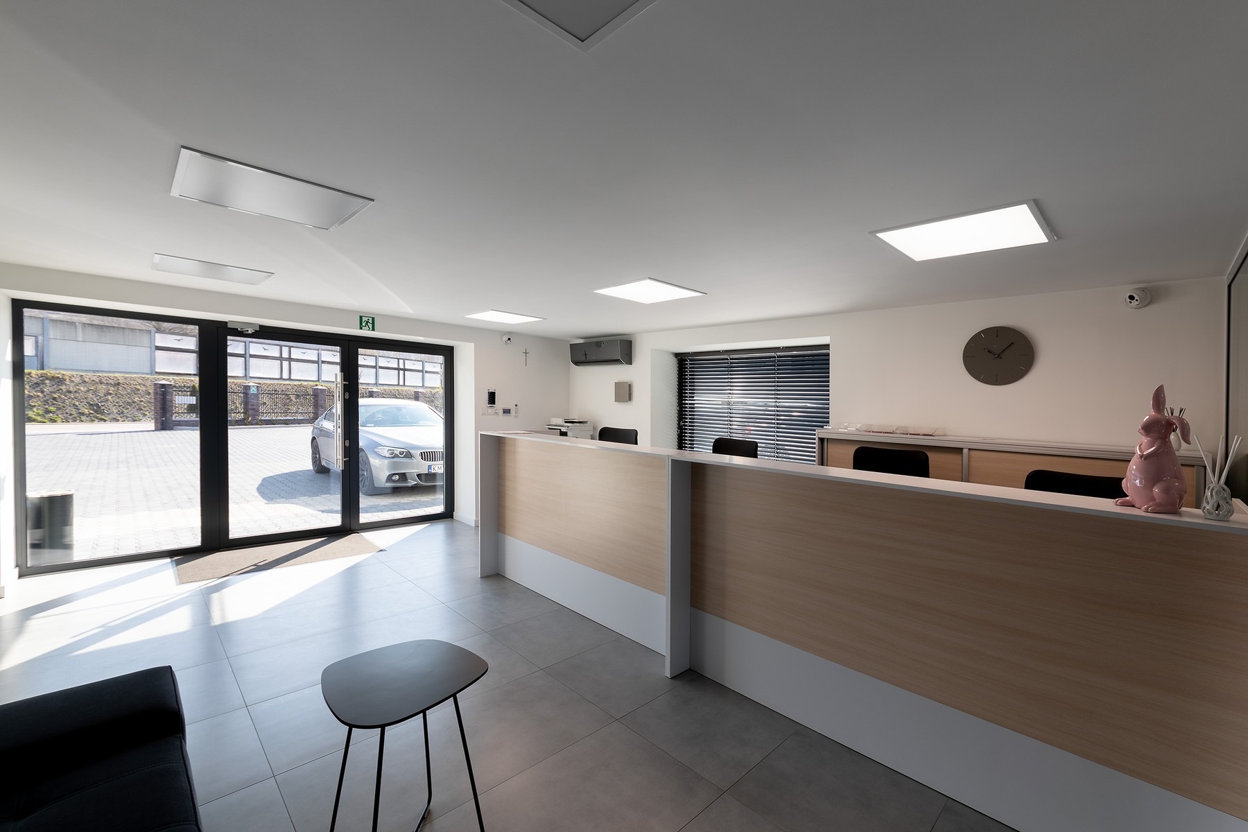 Jasne, minimalistyczne wnętrze recepcji z drewnianym biurkiem, szarą podłogą i widokiem na zewnątrz przez szklane drzwi, gdzie widać zaparkowany samochód; na biurku dekoracyjny różowy królik.