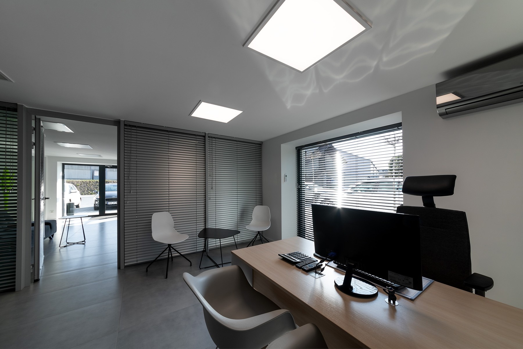 Jasne, minimalistyczne biuro z biurkiem, komputerem, krzesłami i żaluzjami, widok z szerokiego kąta, akcent na czystość i porządek.