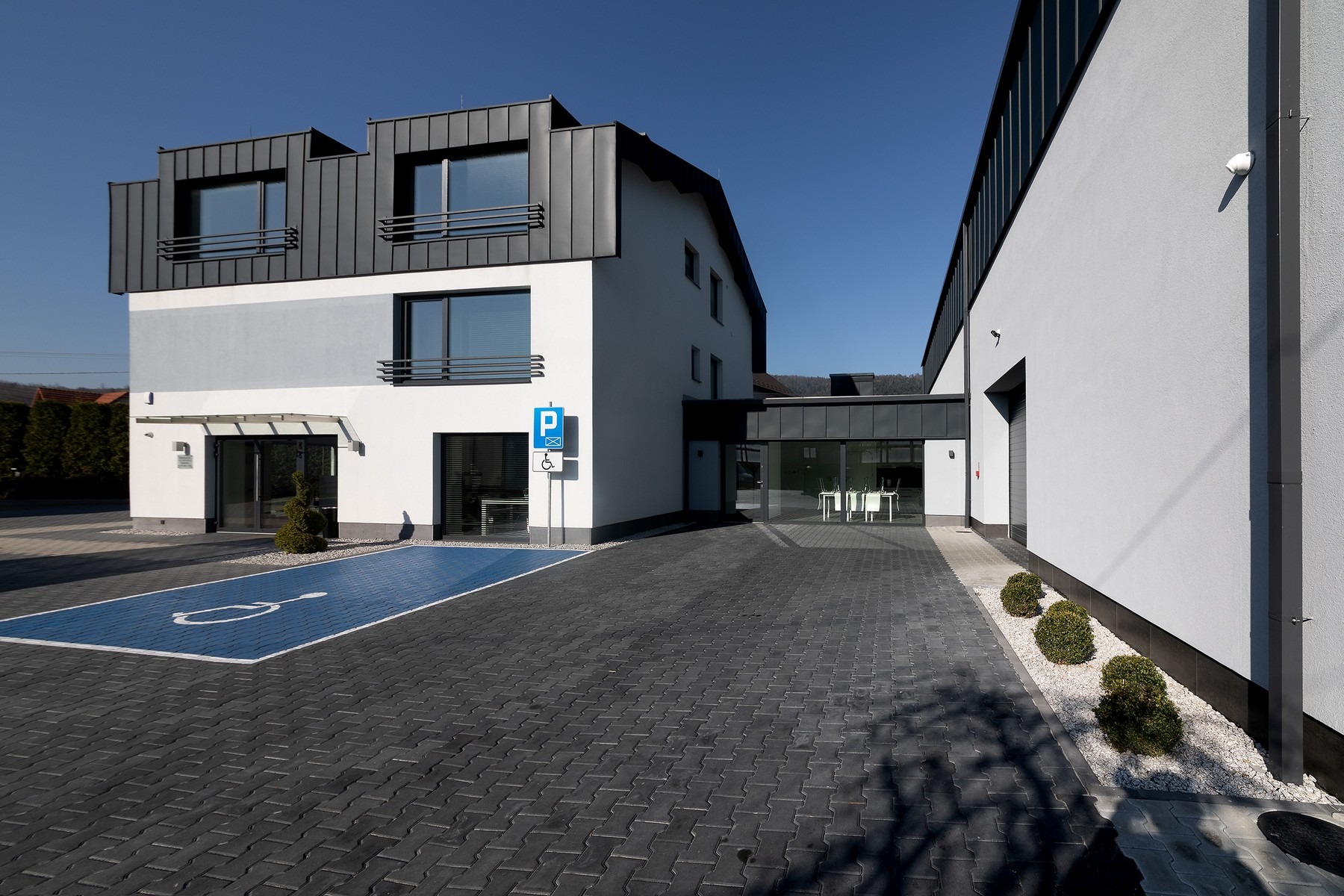 Nowoczesny budynek biurowy z miejscem parkingowym dla niepełnosprawnych oznaczonym niebieską farbą, utrzymany w tonacji bieli i szarości, z elementami metalowymi w kolorze antracytowym i starannie...