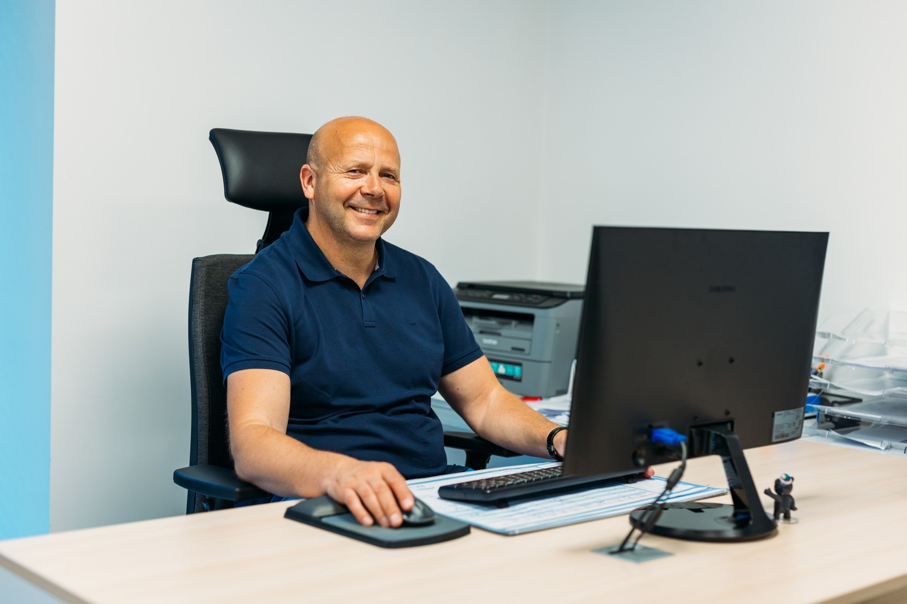 Uśmiechnięty mężczyzna w granatowej koszulce polo siedzi przy biurku z komputerem, za nim drukarka i biało-błękitna ściana.
