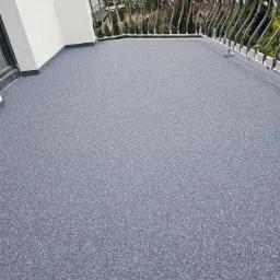 Nowoczesny balkon z szarą, drobnoziarnistą posadzką, wykończony kutą, metalową balustradą z dekoracyjnymi elementami, z widokiem na drzewa w tle.